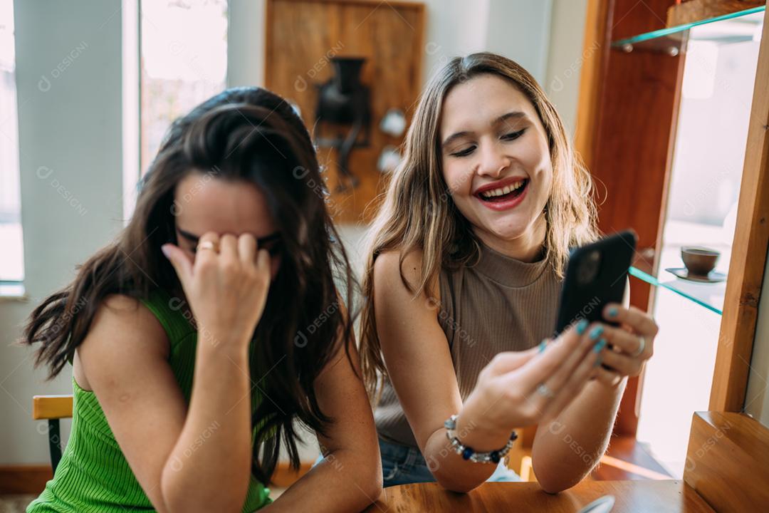 Animadas lindas duas mulheres latinas assistem a um vídeo engraçado no celular moderno, têm expressões