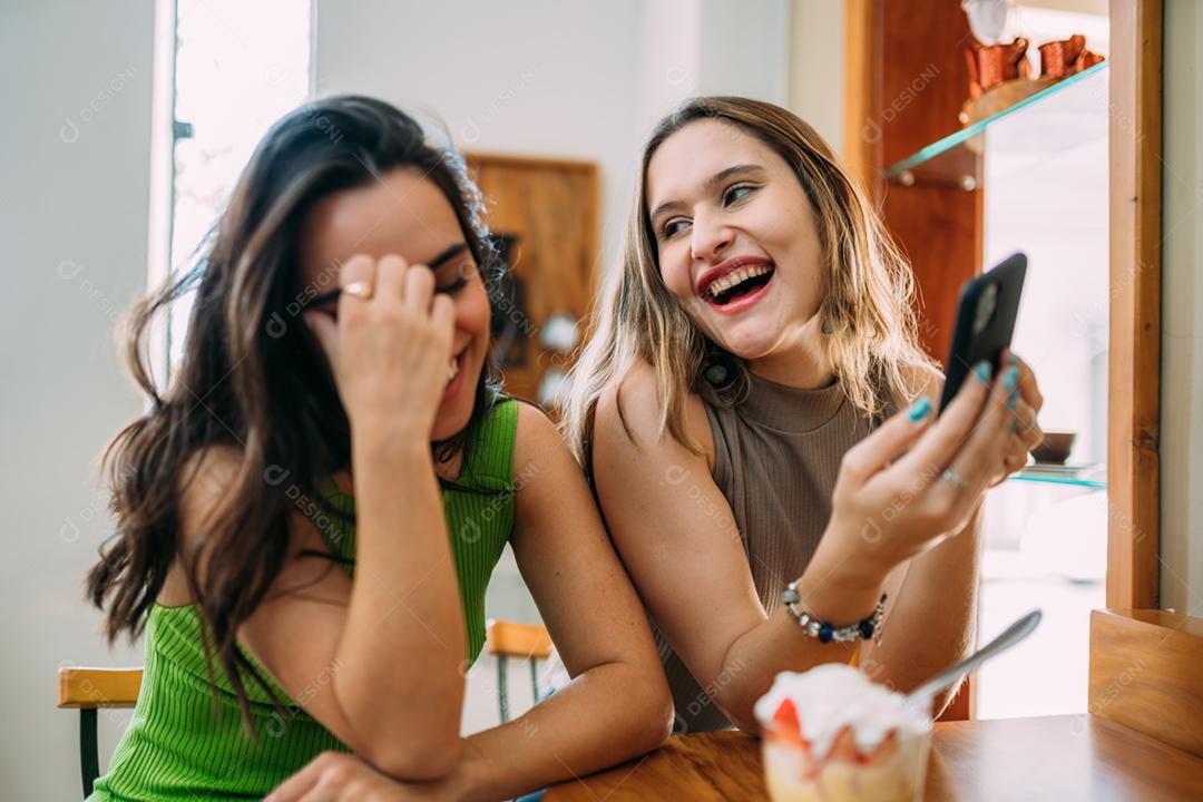 Animadas lindas duas mulheres latinas assistem a um vídeo engraçado no celular moderno, têm expressões