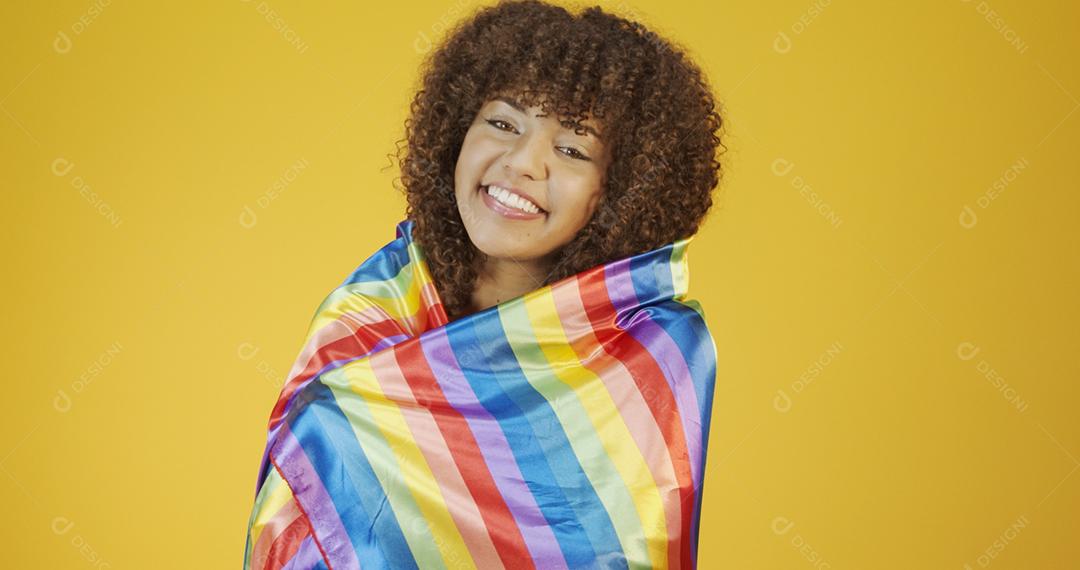 Mulher jovem de cabelo encaracolado cobrindo com a bandeira do orgulho lgbt. Sozinho. Um. Mantendo o punho erguido, cobrindo a bandeira LGBT. Bandeira LGBT+ em fundo amarelo.