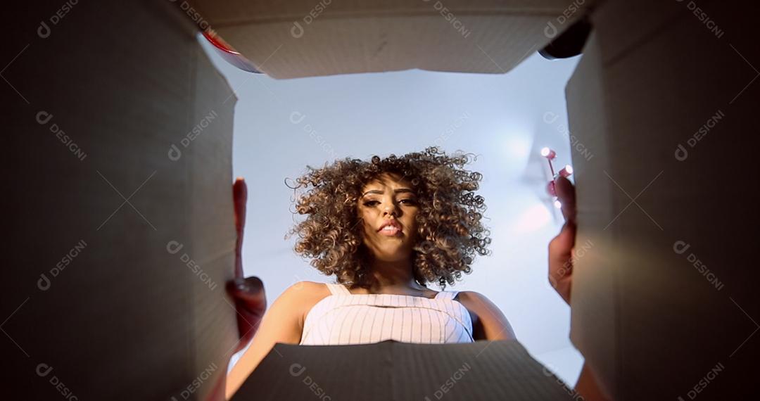 Linda morena com cabelos cacheados abrindo a caixa com seu pedido recebido pelos correios ou empresa de courier. Jovem brasileira recebendo ordem.