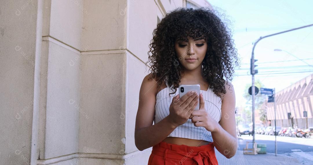 Jovem negra de cabelo encaracolado andando usando o celular. Mensagens de texto na rua. Cidade grande.
