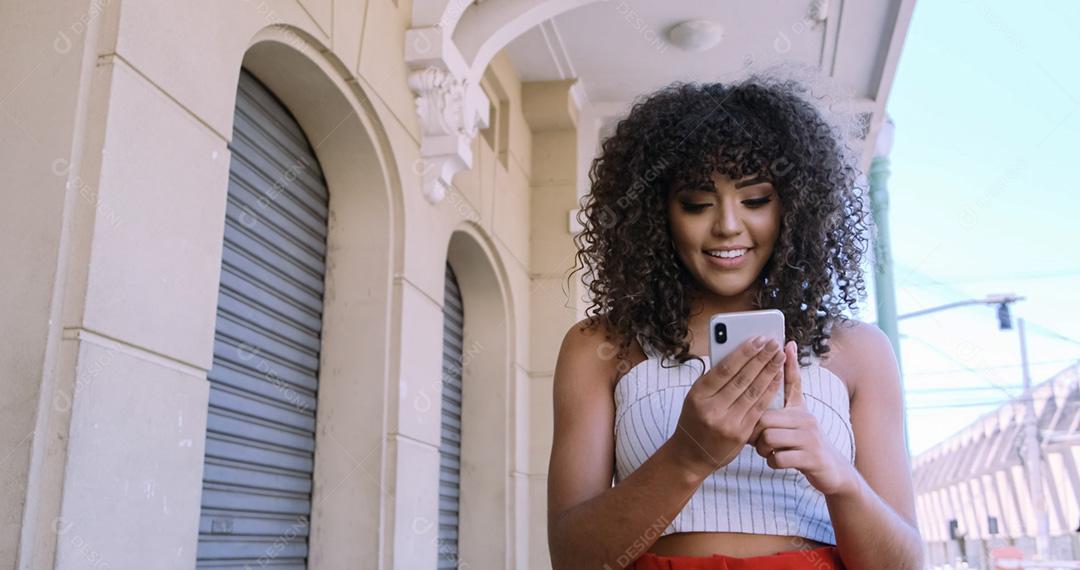 Jovem negra de cabelo encaracolado andando usando o celular. Mensagens de texto na rua. Cidade grande.
