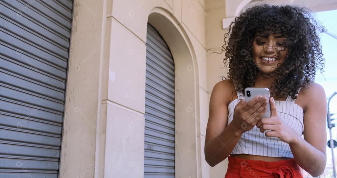 Jovem negra de cabelo encaracolado andando usando o celular. Mensagens de texto na rua. Cidade grande.