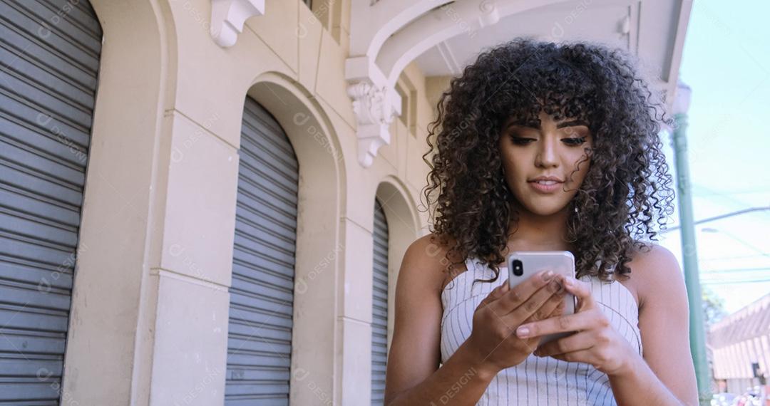 Jovem negra de cabelo encaracolado andando usando o celular. Mensagens de texto na rua. Cidade grande.