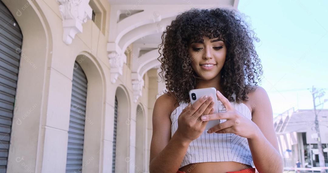 Jovem negra de cabelo encaracolado andando usando o celular. Mensagens de texto na rua. Cidade grande.