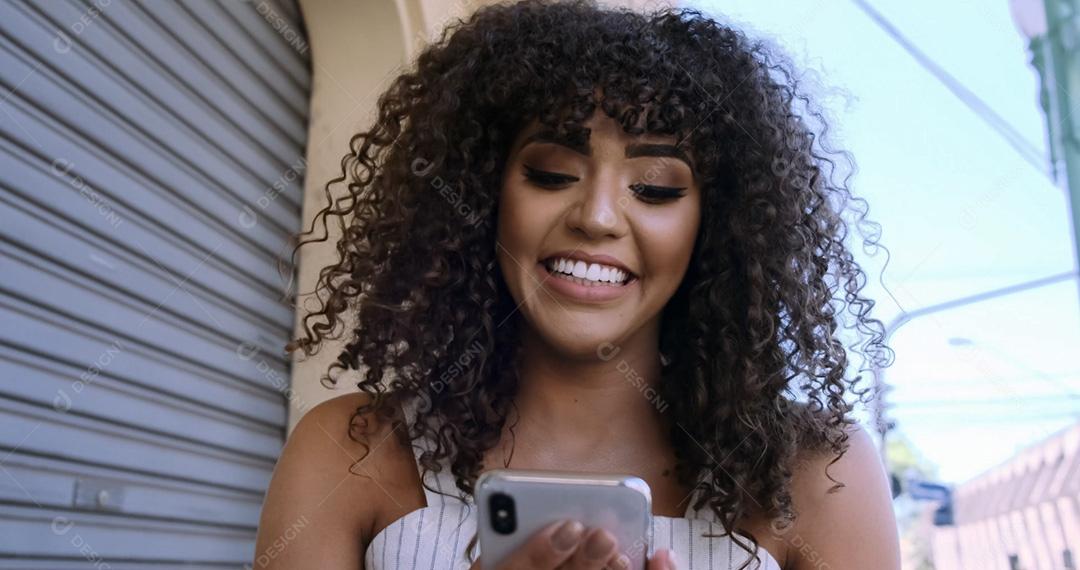 Jovem negra de cabelo encaracolado andando usando o celular. Mensagens de texto na rua. Cidade grande.