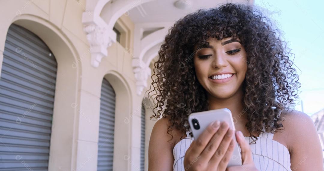 Jovem negra de cabelo encaracolado andando usando o celular. Mensagens de texto na rua. Cidade grande.