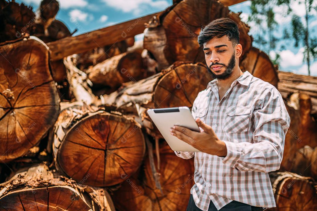 Retrato de jovem latino trabalhando em tablet ao lado de troncos de árvores.
