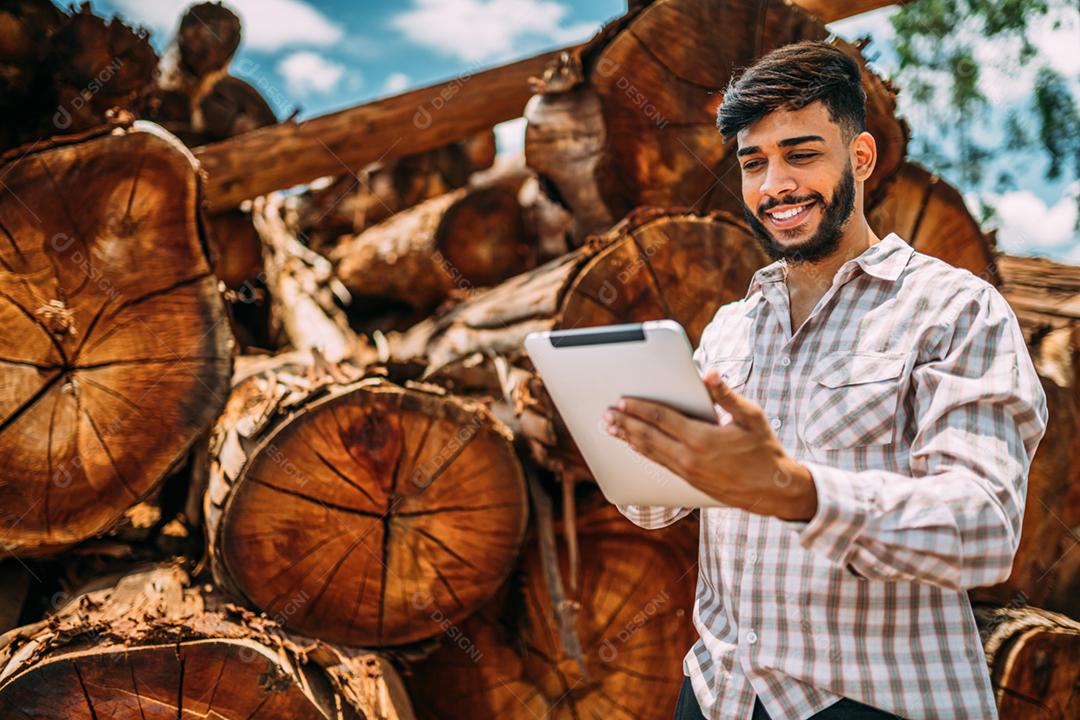 Retrato de jovem latino trabalhando em tablet ao lado de troncos de árvores.