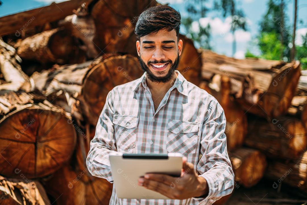 Retrato de jovem latino trabalhando em tablet ao lado de troncos de árvores.