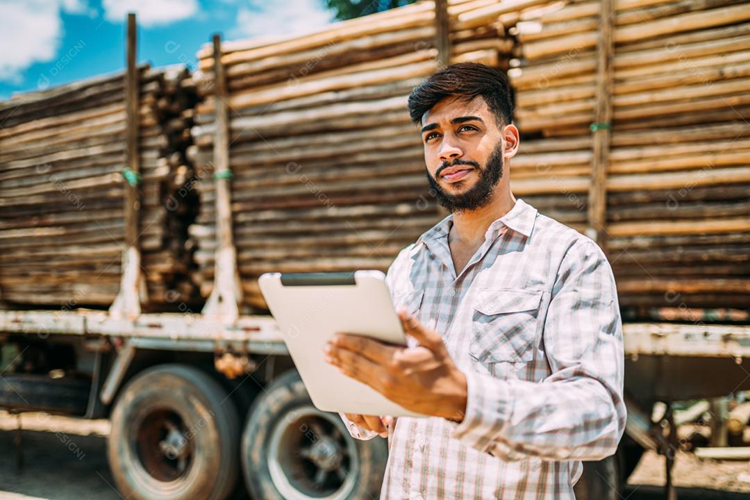 Retrato de jovem latino trabalhando com um tablet digital ao lado de troncos de árvores no caminhão.