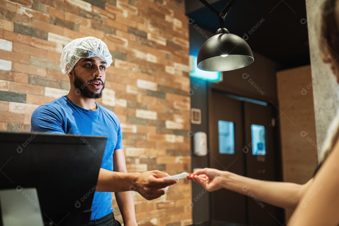 Balconista latina com um boné na cabeça anotando o pedido de uma cliente.