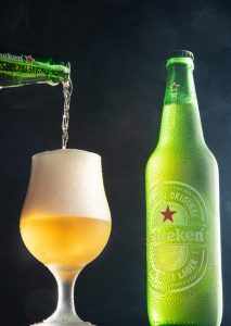 Garrafa e xícara de Heineken envoltas em fumaça fotografadas com fundo escuro