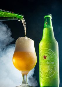 Garrafa e xícara de Heineken envoltas em fumaça fotografadas com fundo escuro
