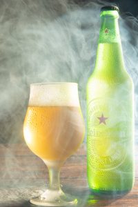 Garrafa e xícara de Heineken envoltas em fumaça fotografadas com fundo escuro