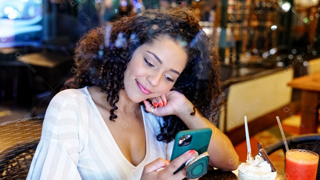 Mulher latina sentada em uma cafeteria com telefone celular tomando café e desviando o olhar.