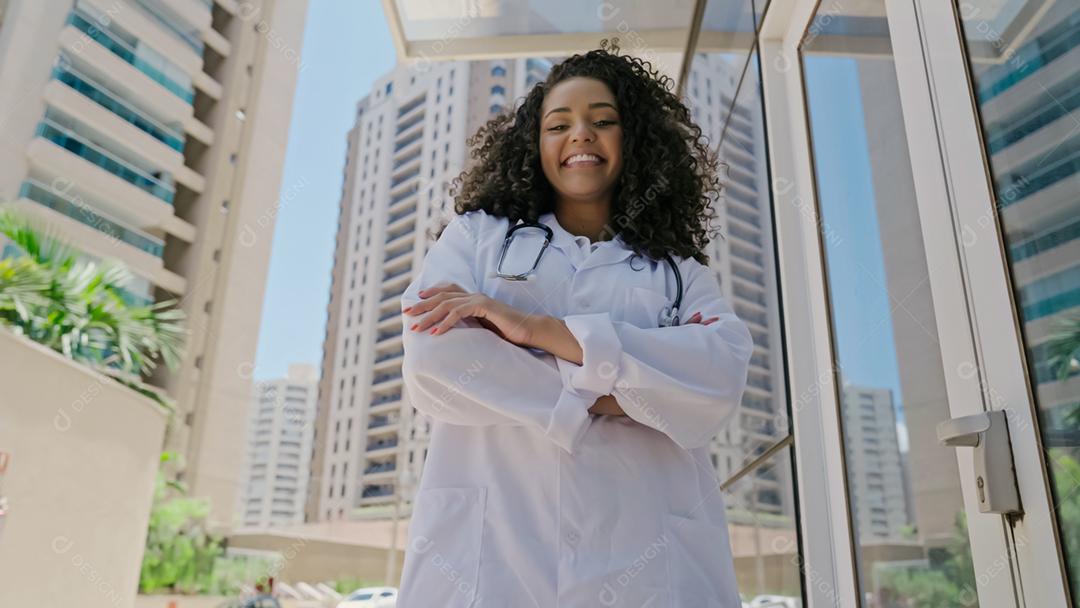 A jovem médica latina usa uniforme branco, jaleco branco, estetoscópio e olhando para a câmera