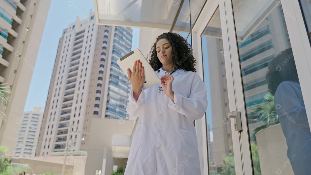 Jovem médica latina usa uniforme branco, usando tablet digital no hospital