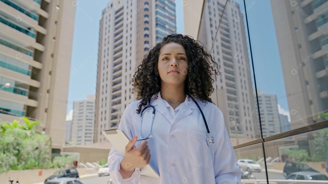 Jovem médica latina usa uniforme branco, usando tablet digital no hospital