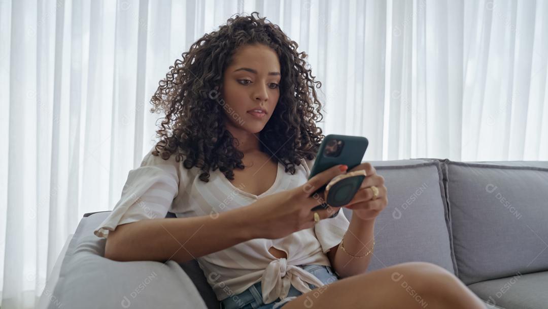 Feliz dama latina relaxa em casa sozinha, sente-se na sala e compartilhe boas notícias nas mídias sociais via celular.