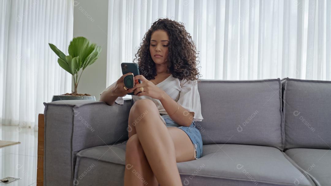 Feliz dama latina relaxa em casa sozinha, sente-se na sala e compartilhe boas notícias nas mídias sociais via celular.