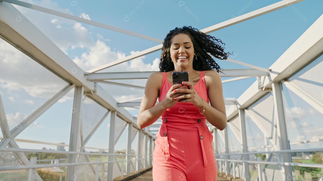 Jovem mulher latina de cabelo encaracolado andando usando o celular. Mensagens de texto na rua. Cidade grande.