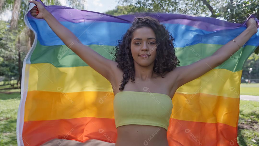 Mulher jovem de cabelo encaracolado cobrindo com a bandeira do orgulho lgbt. Mantendo o punho erguido, cobrindo a bandeira LGBT +