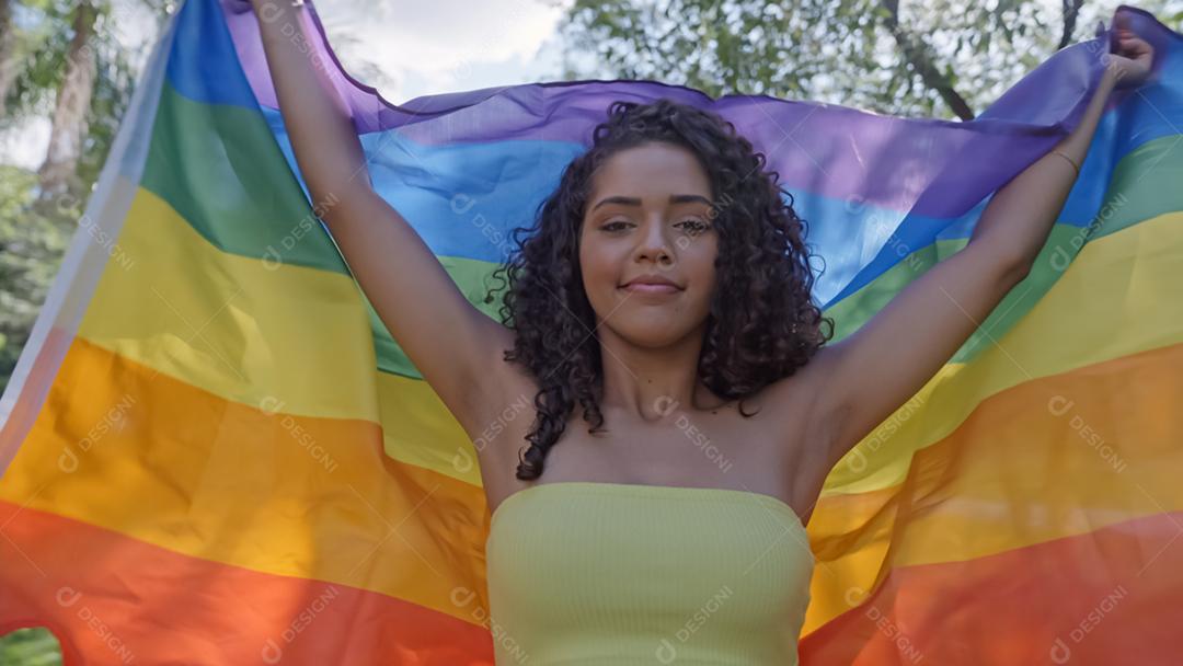 Mulher jovem de cabelo encaracolado cobrindo com a bandeira do orgulho lgbt. Mantendo o punho erguido, cobrindo a bandeira LGBT +