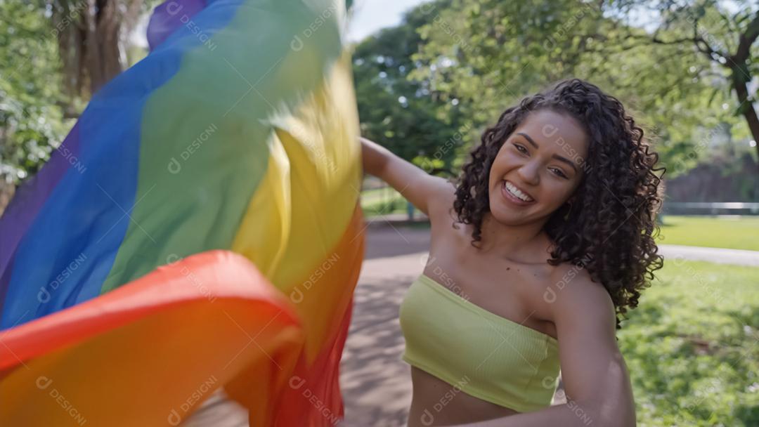 Mulher jovem de cabelo encaracolado cobrindo com a bandeira do orgulho lgbt. Mantendo o punho erguido, cobrindo a bandeira LGBT +