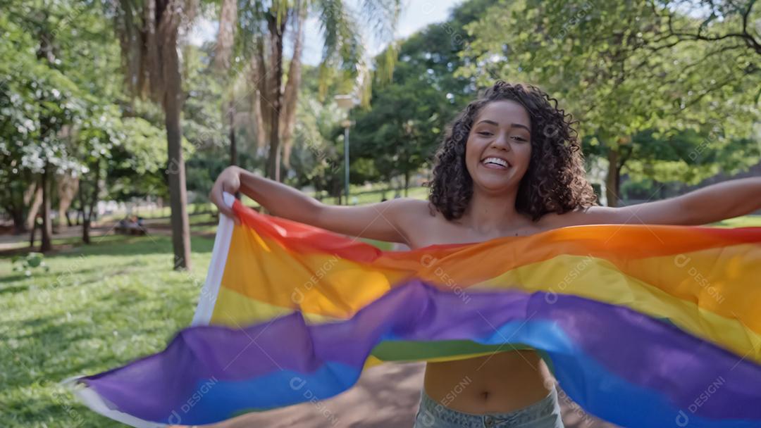 Mulher jovem de cabelo encaracolado cobrindo com a bandeira do orgulho lgbt. Mantendo o punho erguido, cobrindo a bandeira LGBT +