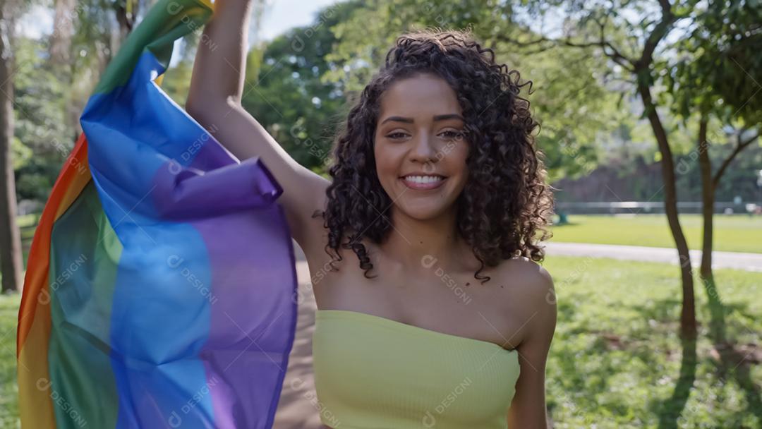 Mulher jovem de cabelo encaracolado cobrindo com a bandeira do orgulho lgbt. Mantendo o punho erguido, cobrindo a bandeira LGBT +