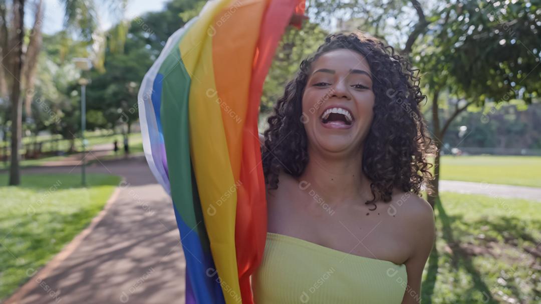 Mulher jovem de cabelo encaracolado cobrindo com a bandeira do orgulho lgbt. Mantendo o punho erguido, cobrindo a bandeira LGBT +