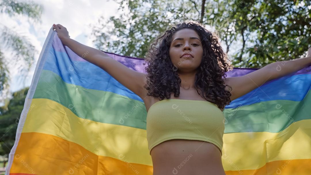 Mulher jovem de cabelo encaracolado cobrindo com a bandeira do orgulho lgbt. Mantendo o punho erguido, cobrindo a bandeira LGBT +