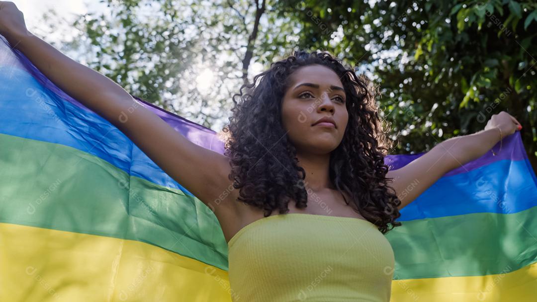 Mulher jovem de cabelo encaracolado cobrindo com a bandeira do orgulho lgbt. Mantendo o punho erguido, cobrindo a bandeira LGBT +
