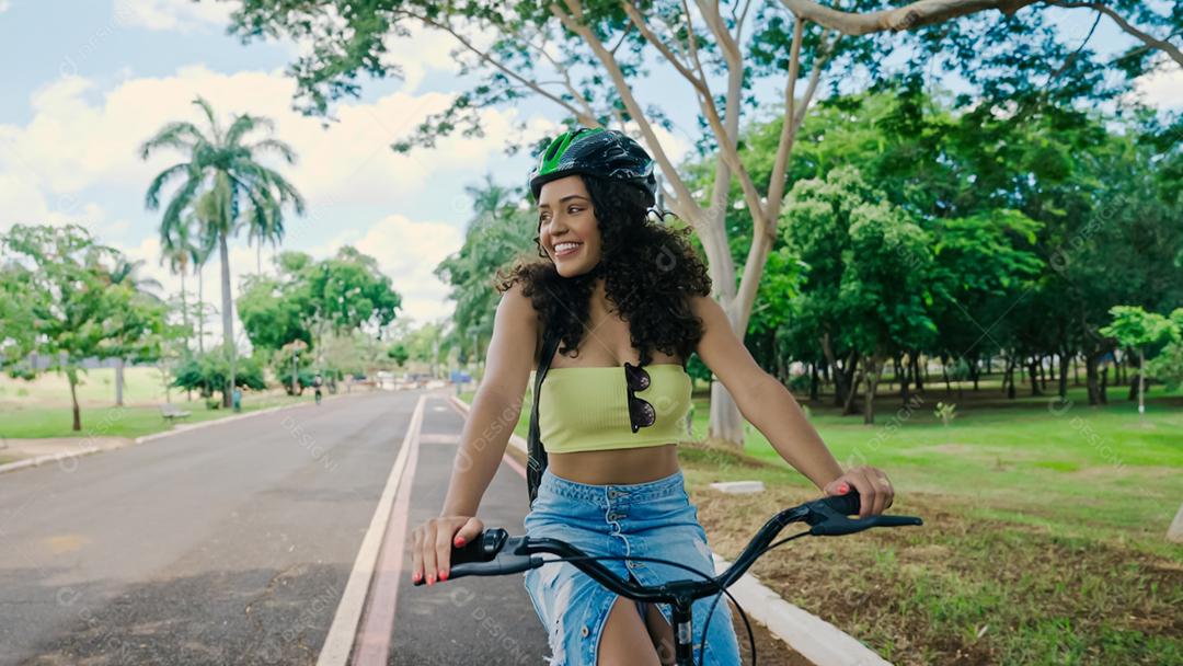 Jovem mulher latina em capacete protetor está andando de bicicleta ao longo da ciclovia em um parque da cidade