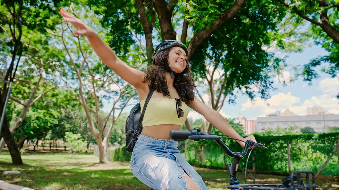 Jovem mulher latina em capacete protetor está andando de bicicleta ao longo da ciclovia em um parque da cidade
