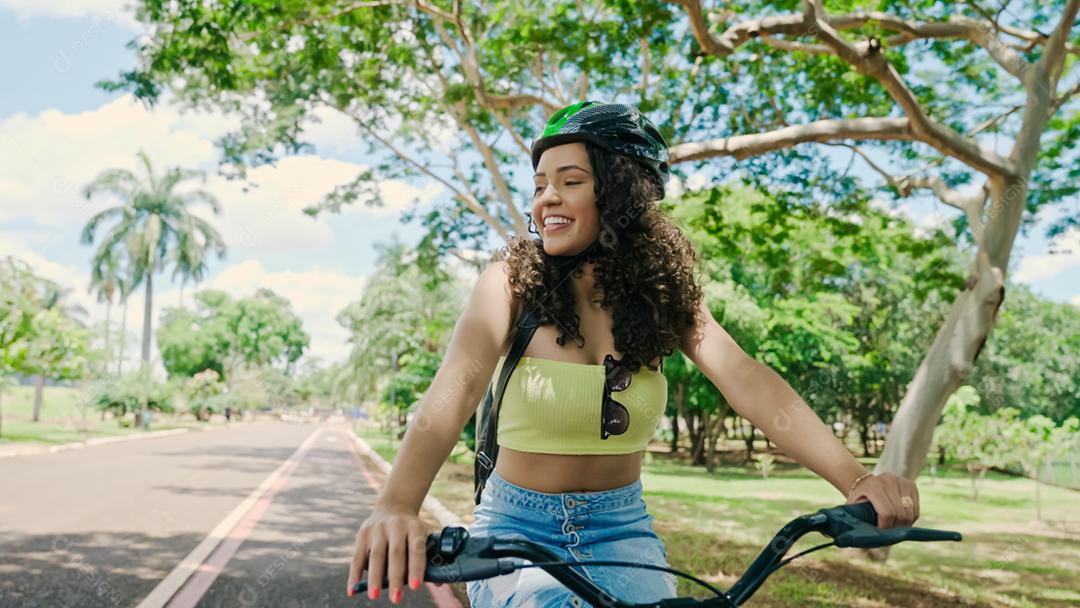Jovem mulher latina em capacete protetor está andando de bicicleta ao longo da ciclovia em um parque da cidade