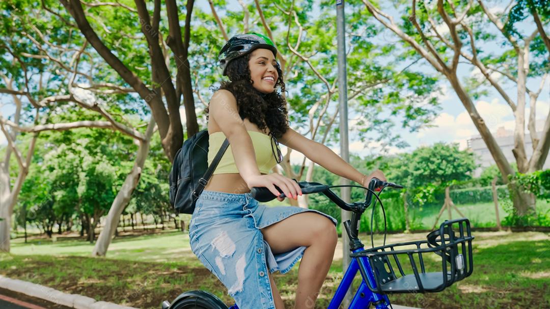 Jovem mulher latina em capacete protetor está andando de bicicleta ao longo da ciclovia em um parque da cidade