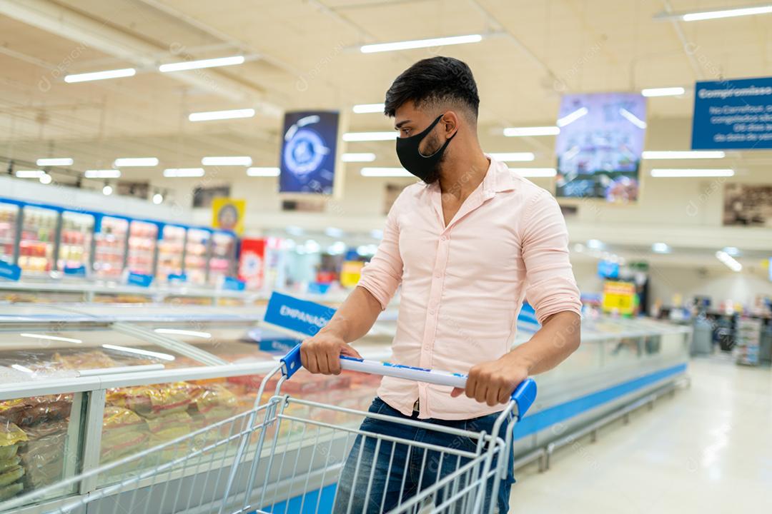 Homem latino-americano com carrinho de compras em um supermercado, usando máscara protetora