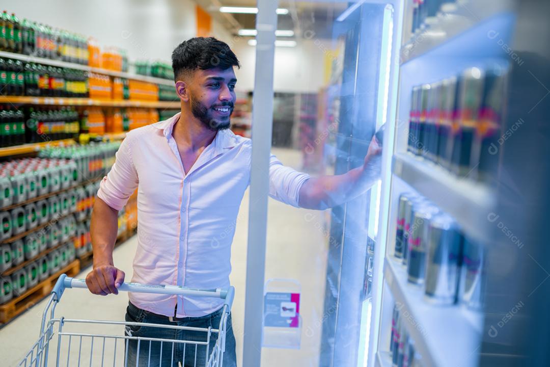Um homem latino-americano com um carrinho de compras tira comida congelada da geladeira do supermercado
