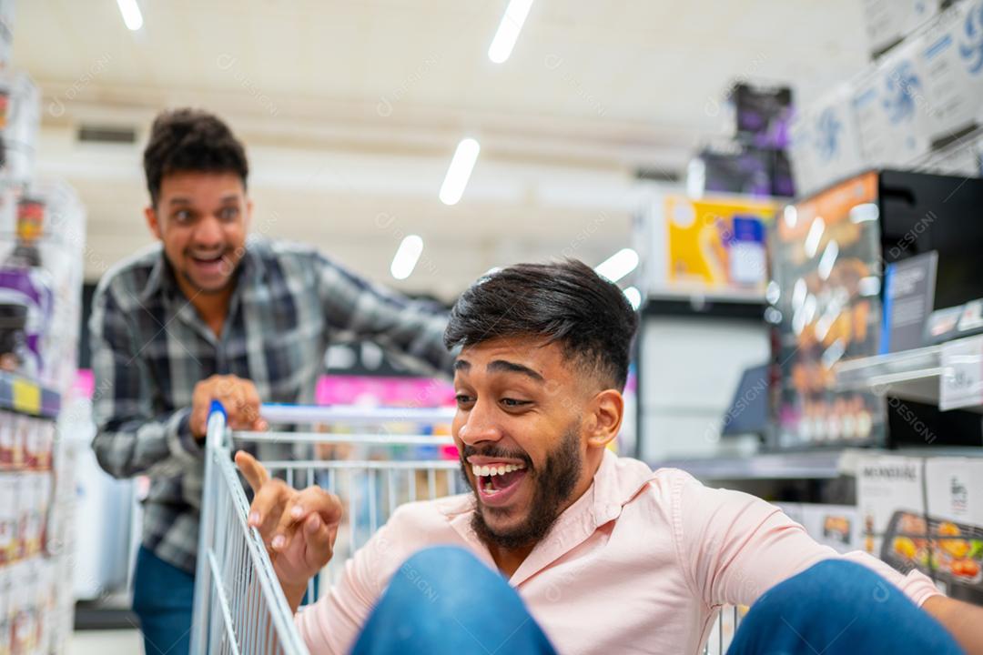 No supermercado: homem empurra o carrinho de compras com um amigo sentado nele. Felizes amigos latino-americanos