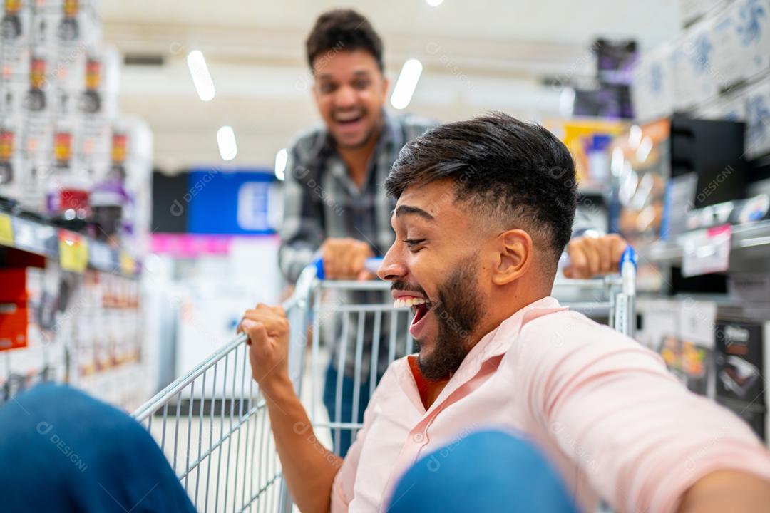 No supermercado: homem empurra o carrinho de compras com um amigo sentado nele. Felizes amigos latino-americanos