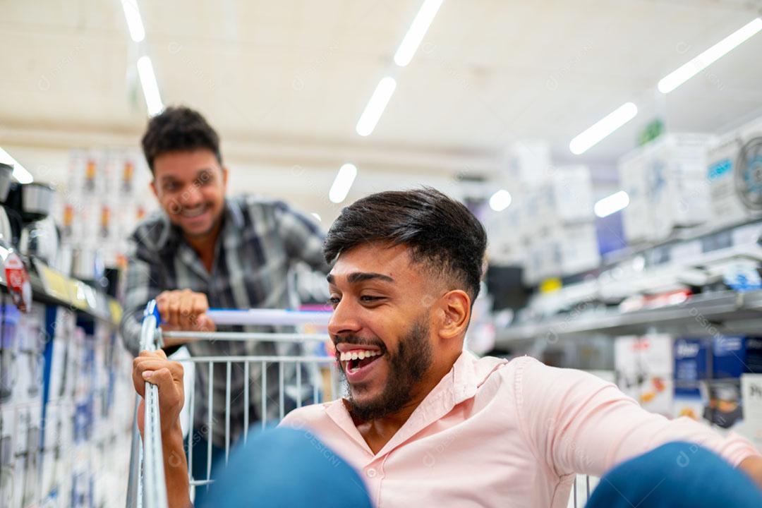 No supermercado: homem empurra o carrinho de compras com um amigo sentado nele. Felizes amigos latino-americanos