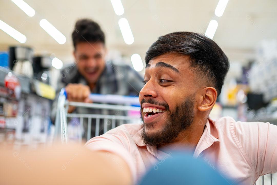 No supermercado: homem empurra o carrinho de compras com um amigo sentado nele. Felizes amigos latino-americanos