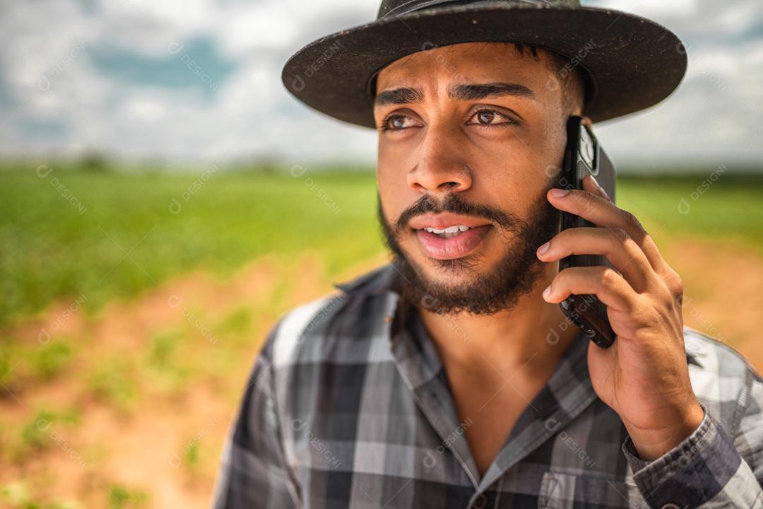 Agricultor latino-americano trabalhando na plantação. Jovem falando no celular, usando chapéu
