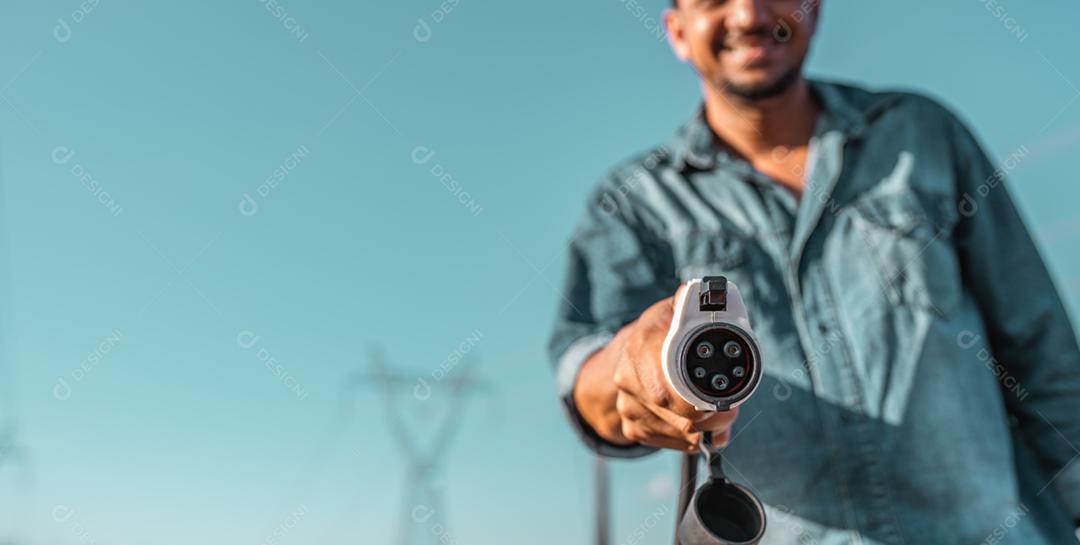 A mão humana está segurando o carregamento do carro elétrico conectado ao carro elétrico. Torres de alta tensão ao fundo.
