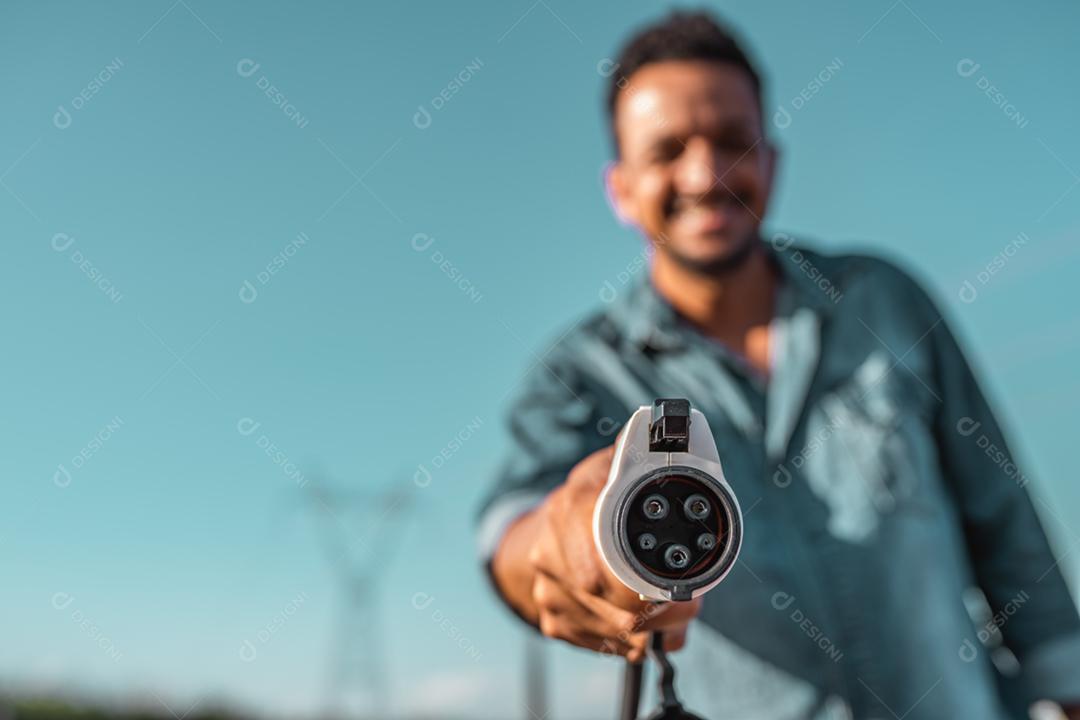 A mão humana está segurando o carregamento do carro elétrico conectado ao carro elétrico. Torres de alta tensão ao fundo.