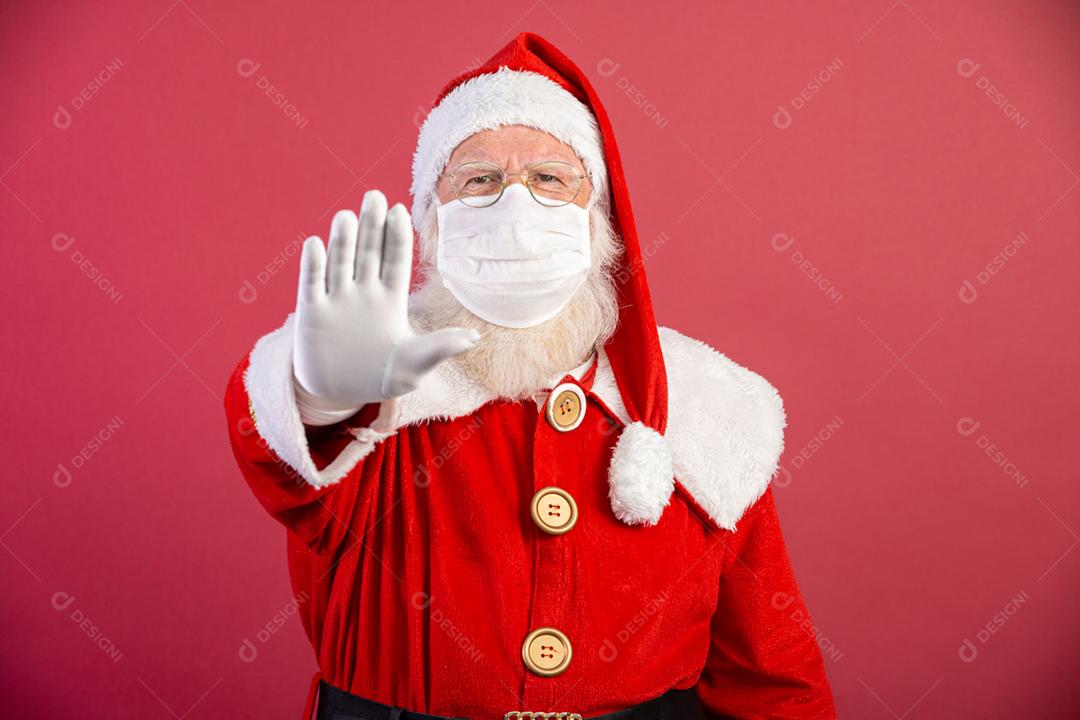 Verdadeiro Papai Noel em fundo vermelho, usando máscara protetora e com a mão aberta na frente