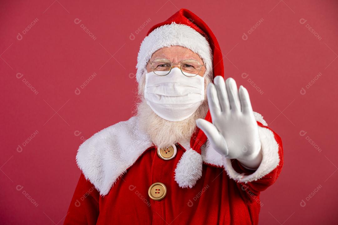 Verdadeiro Papai Noel em fundo vermelho, usando máscara protetora e com a mão aberta na frente