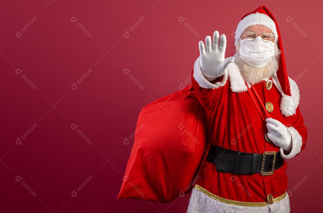 Verdadeiro Papai Noel em fundo vermelho, usando máscara protetora e com a mão aberta na frente